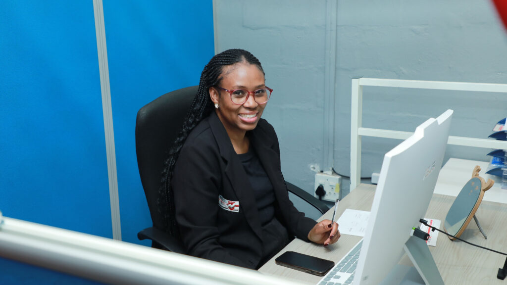 A woman with long braids and glasses sits at a desk, smiling at the camera. She is wearing a black blazer and working on a computer in a modern office setting. A smartphone lies on the desk beside her.